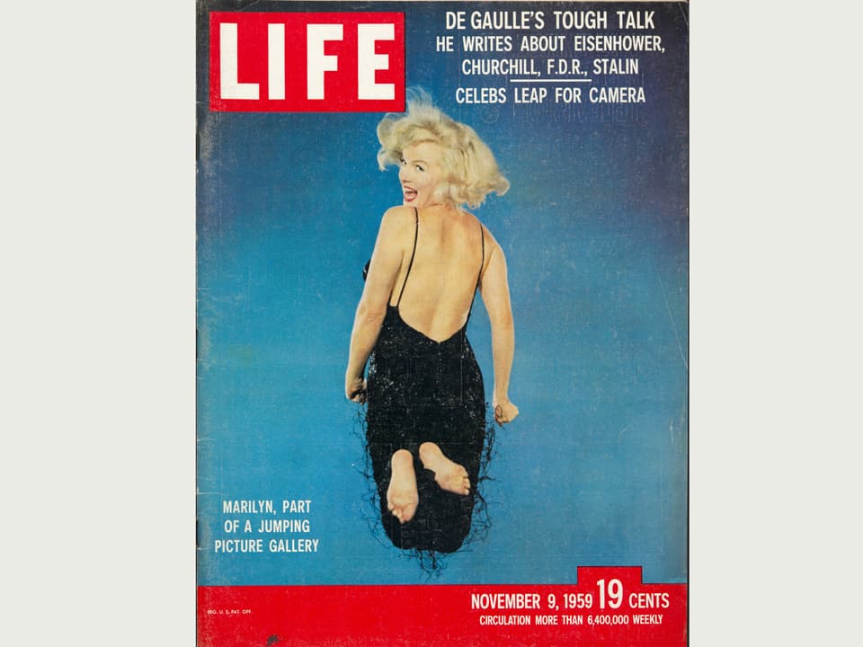 Marilyn Monroe, barfuss, in schwarzem, rückenfreien Kleid springt in die Höhe und lacht mit rückwärts gewandtem Kopf in die Kamera.