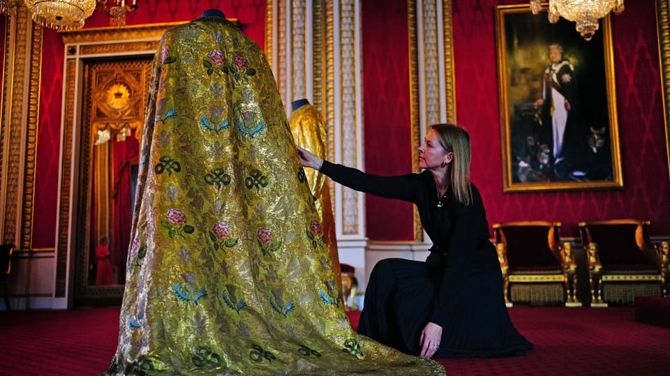 Foto eines dunkelrot-samtenen Raums, in der Mitte ein goldener Mantel mit Stickereien aufgestellt, Frau richtet ihn.