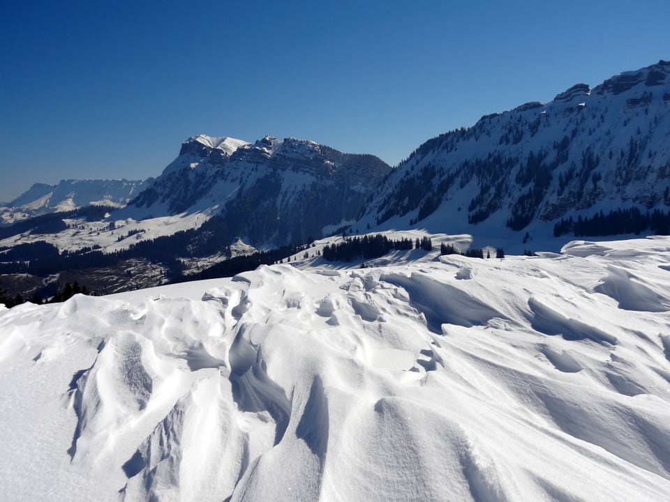 Aufnahme in Richtung Hohgant (Kanton Bern),  im Hintergrund bilden Schneeverwehungen eine wellige Schneeoberfläche, darüber blauer Himmel lauernden versteckten Stellen mit verfrachtetem Schnee 