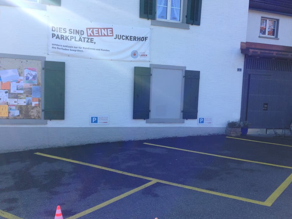 Ein Haus, davor Parkplätze, an der Mauer ein Plakat mit der Aufschrift "Dies sind keine Juckerhof Parkplätze!"