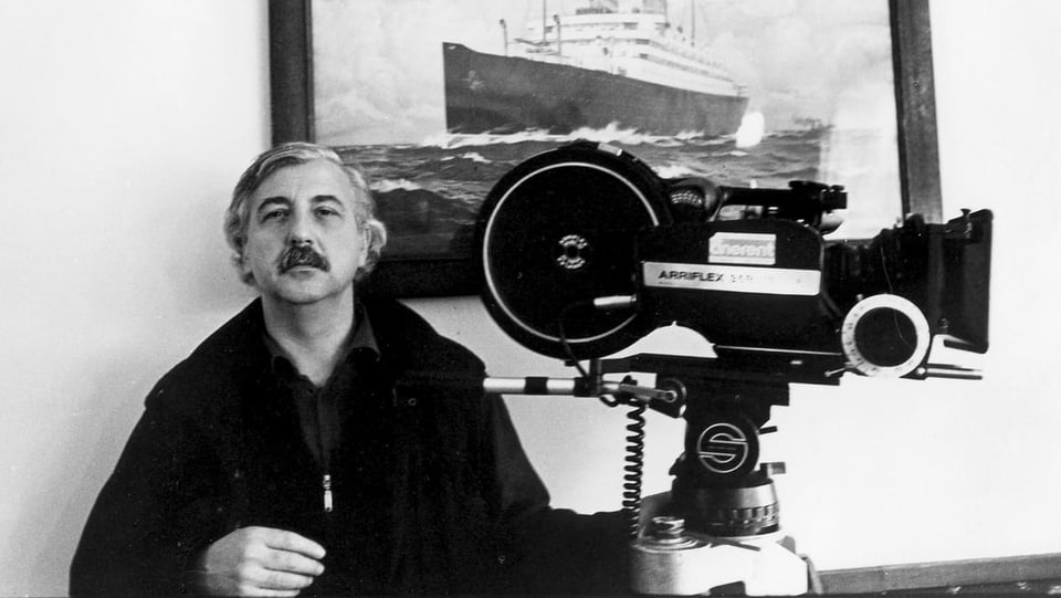 Ein Mann mit Schnauz sitzt neben einer grossen Kamera vor einem Bild mit einem Schiff.