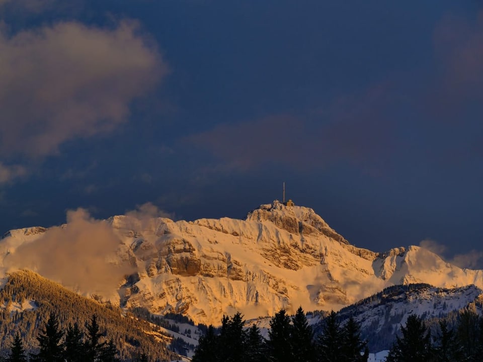 Verschneiter Berggipfel im Sonnenuntergang mit Wolken.