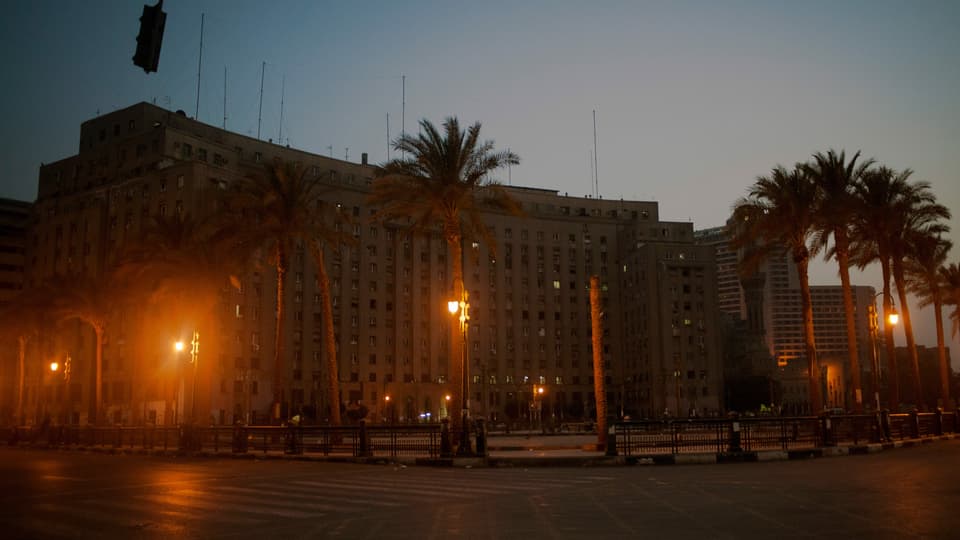 Ein grosses Gebäude bei Nacht steht am Rande eines Platzes neben ein paar Palmen.