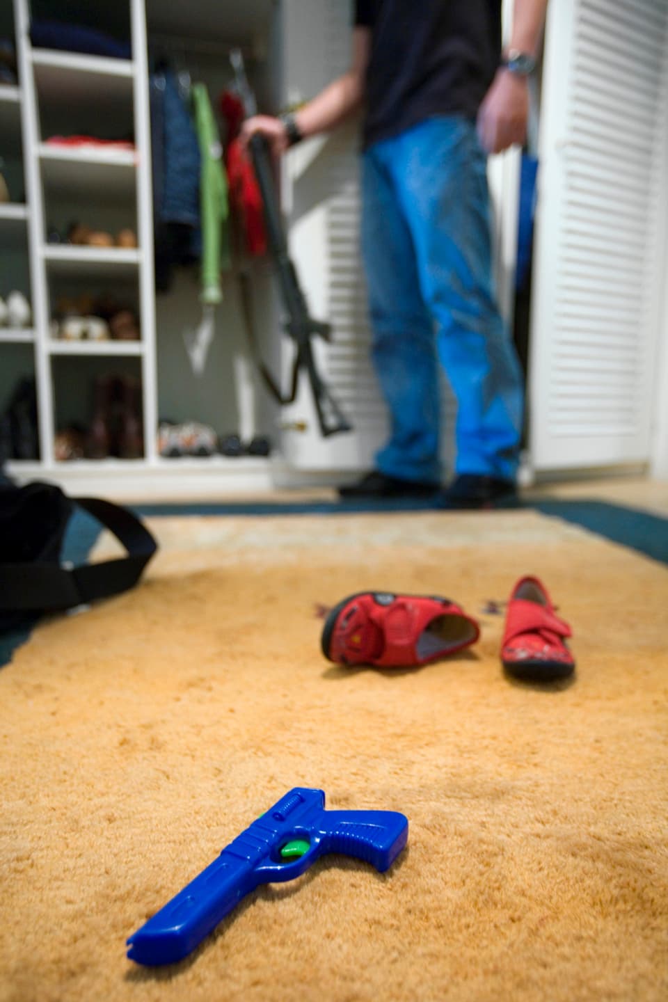 Ein Mann stellt sein Sturmgewehr in den Kleiderschrank, im Vordergrund liegen Kinderschuhe und eine Spielzeugpistole (2006).