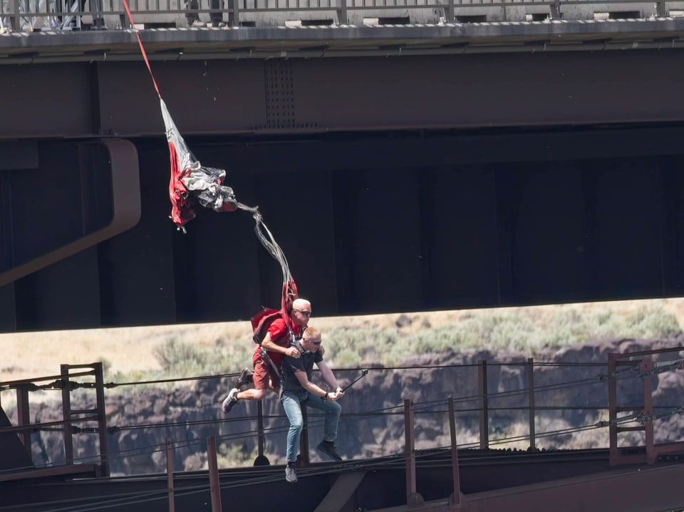 ZWei Menschen springen gemeinsam, aneinandergebunden, von einer Brücke, danach öffnen sie einen Fallschirm.