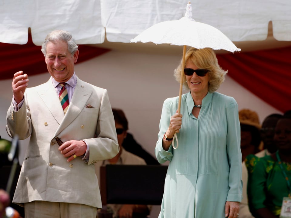 Ein Mann im Anzug geht neben einer Frau im blauen Kleid und einen Sonnenschirm haltend.