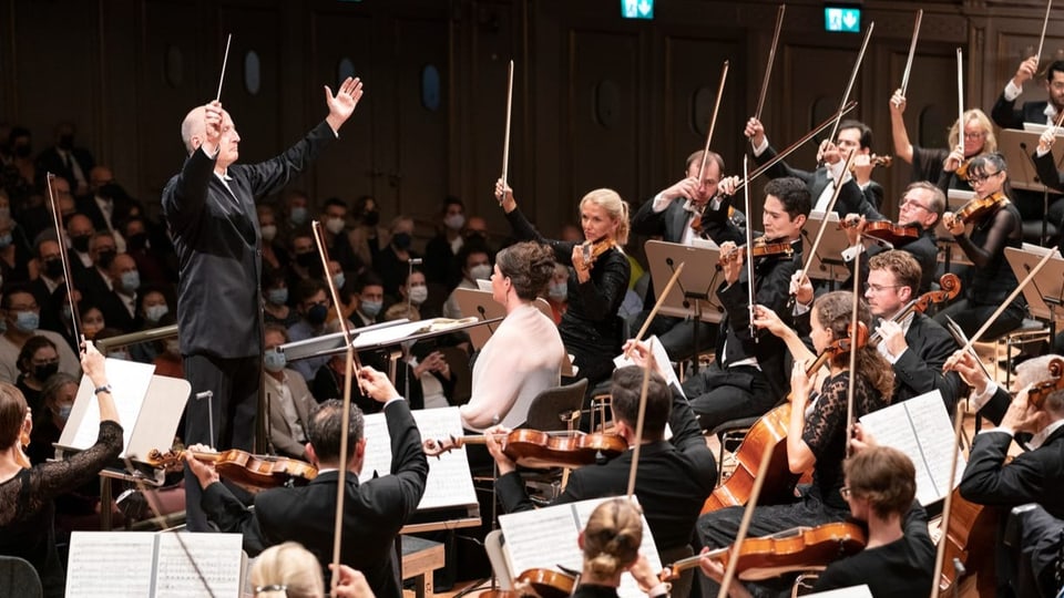 Link älterer Herr im Anzug, schwingt Dirigentenstab. Rechts sitzende Musiker mit Sreichinstrumenten.