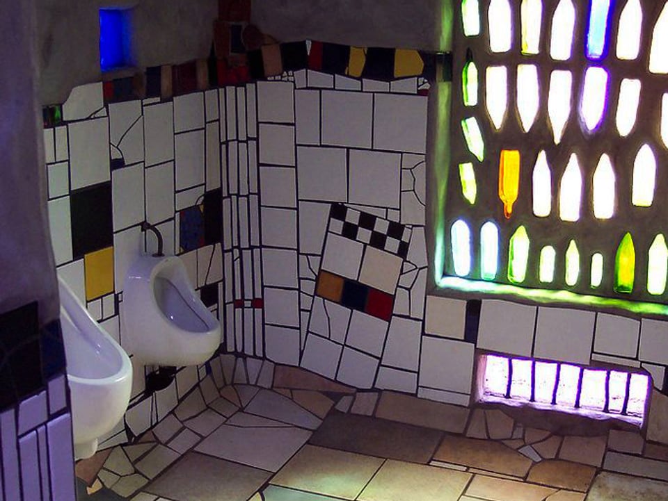 Toilette mit zwei Pissoirs mit bunten Wänden und einer Wand, in die ganze Flaschen eingelassen sind.