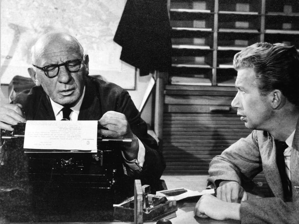 Ein älterer Mann sitzt an einer Schreibmaschine. Heinz sitzt daneben und schau ihn an.