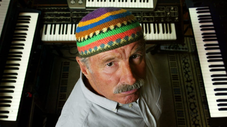 Blick von oben auf einen Mann mit einer bunten Mütze, der vor mehreren Keyboards steht und von unten in die Kamera schaut.