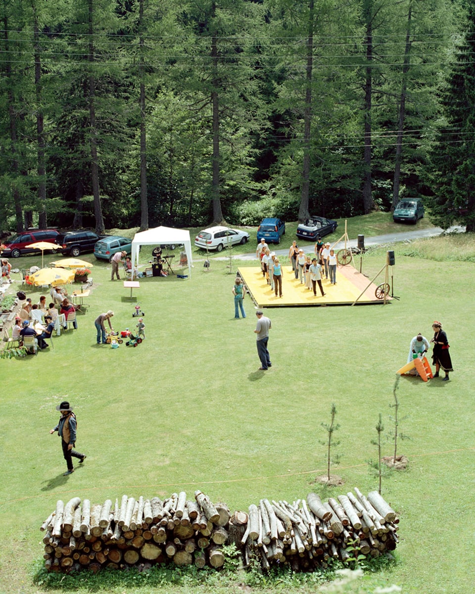 Auf einer grünen Wiese am Waldrand steht eine Holzbühne, auf der eine Grupe von Menschen tanzt.
