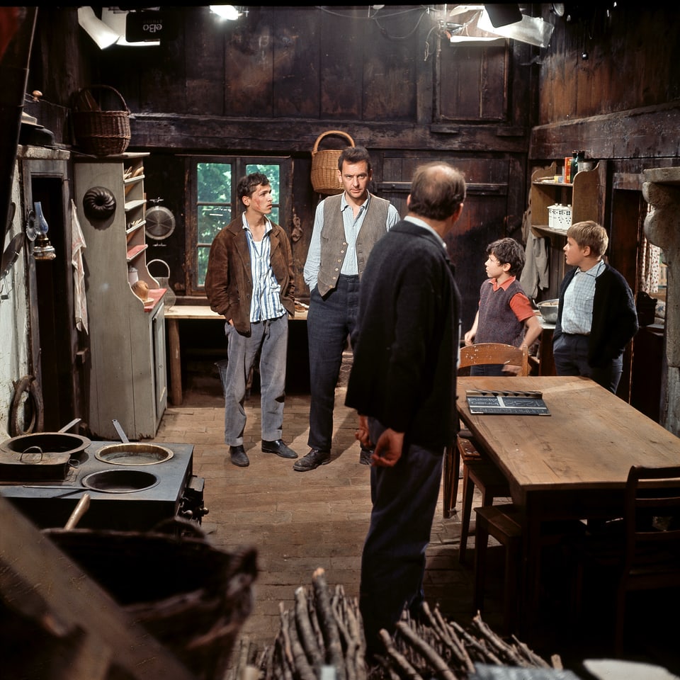 Drei Männer und zwei Buben stehen in einer Küche. Vorne Links der Herd, rechts der Esstisch. An der Decke hängen Filmscheinwerfer.