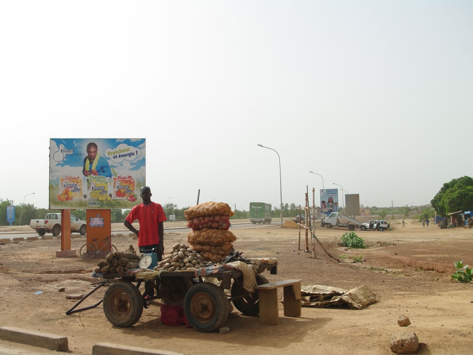 Ein Mann steht vor einem Wagen, auf dem er Kartoffeln und andere Nahrungsmittel feilhält. Im Hintergrund ist ein grosses Plakat zu sehen, das für ein Erfrischungsgetränk wirbt.