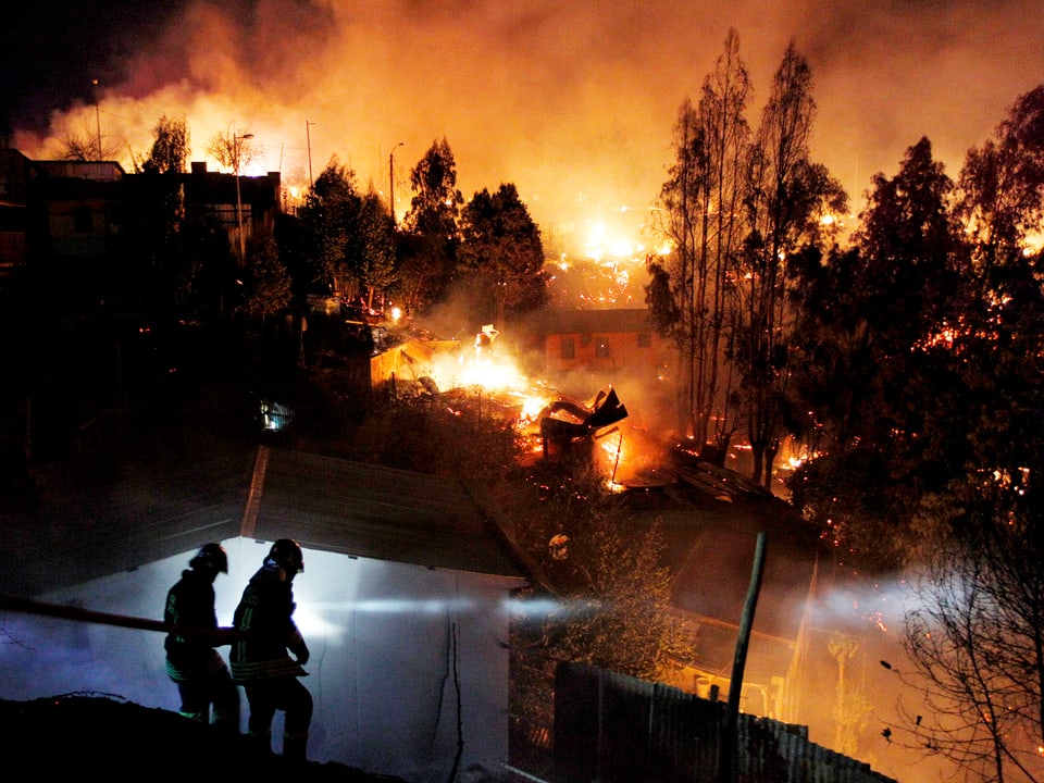 Feuerwehrmänner mit Löschschlauch. Im Hintergrund wütet der Brand.