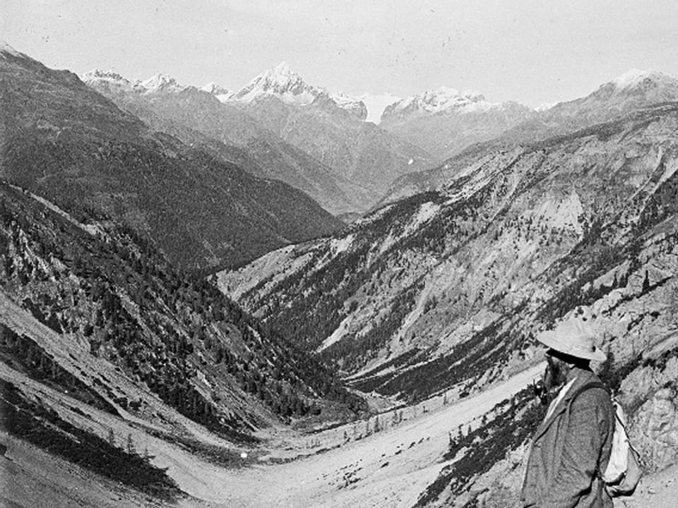 Ein Tal, von oben gesehen, am unteren Bildrand ein Mann in Wanderkleidung mit Hut.