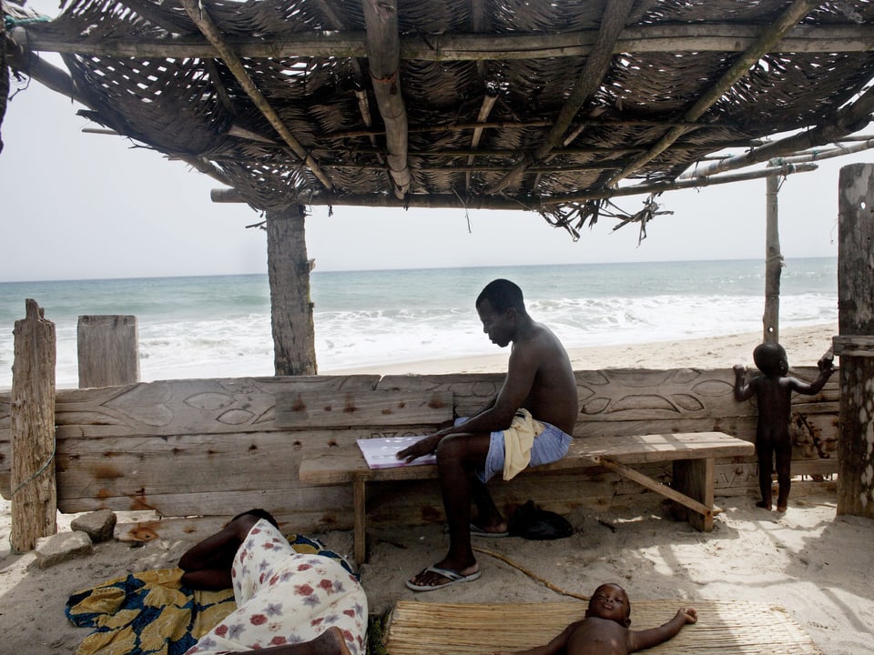 Eine Familie in einem Verschlag mit Dach am Strand. Die Frau und ein kleines Kind liegen auf Matten im Sand. Der Mann sitzt auf einer Bank und liest.