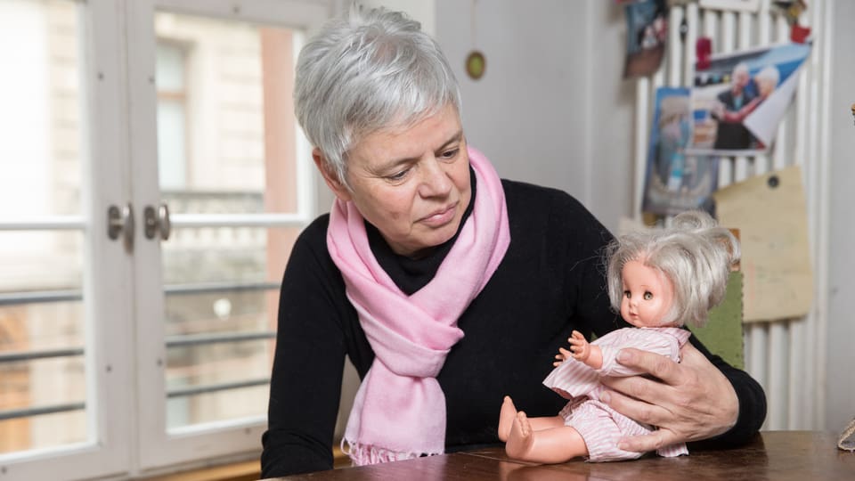 Eine Frau sitzt an einem Tisch. Sie schaut auf eine Puppe, die sie in der Hand hält. 