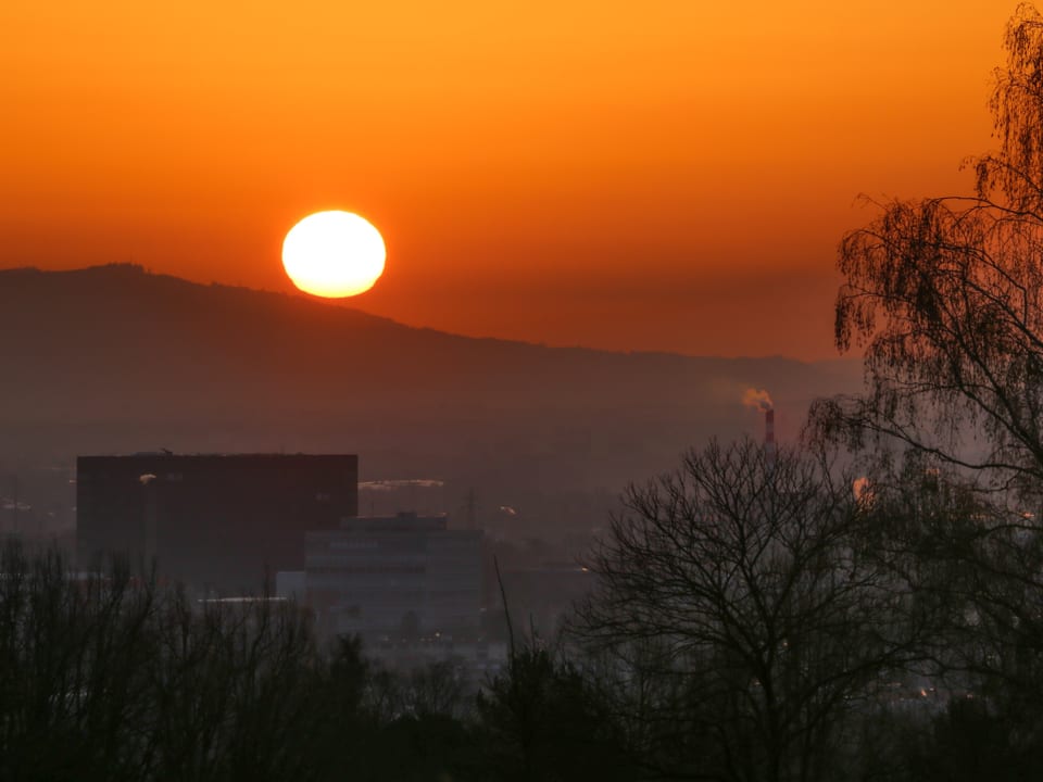 Morgensonne knapp über dem Hügel färbt Himmel gelb-orange, im Vordergrund Siedlung. 