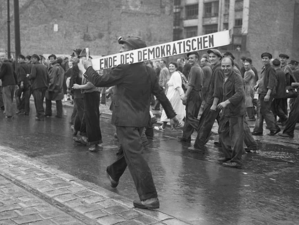 Schwarzweiss-Bild: Mann trägt Schild mit der Aufschrift «Ende des Demokratischen» auf den Schultern durch die Strassen.