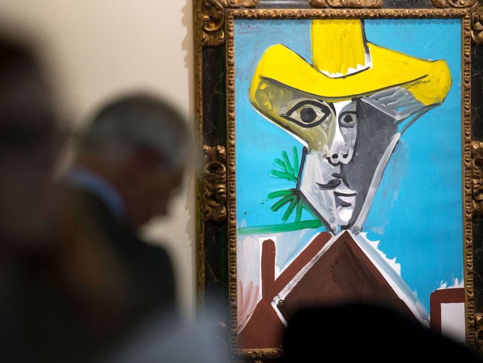 Ein Mann steht vor einem Bild eines abstrakten Kopfes mit gelbem Sombrero, vor blauem Hintergrund.
