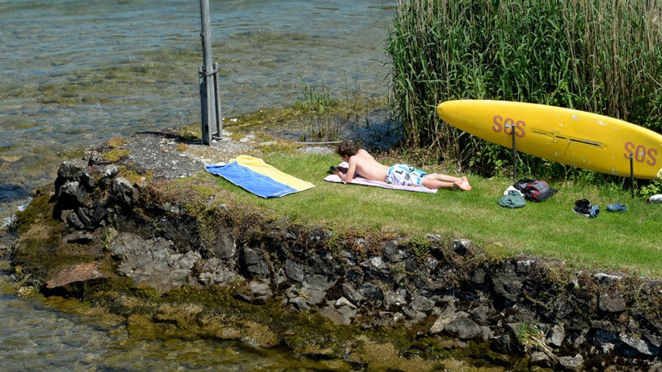 Ein Mann liegt auf einer Wiese am See in der Sonne.