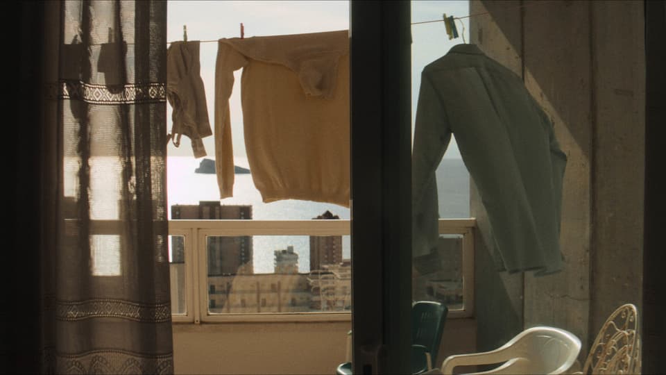 Ausblick von einem Balkon auf das Meer und Gebäude. Dazwischen hängen Kleidungsstücke auf einer Wäscheleine.