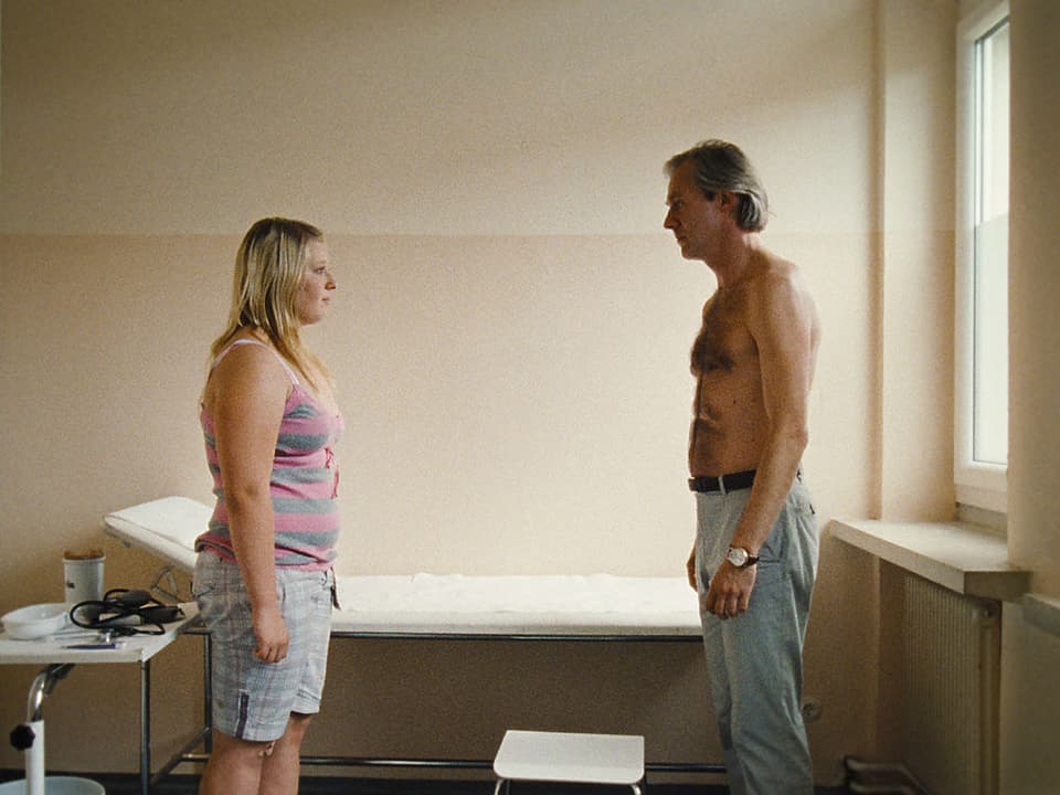 Melanie und der Arzt ((Joseph Lorenz) stehen sich im Behandlungszimmer gegenüber, er mit entblösstem Oberkörper.