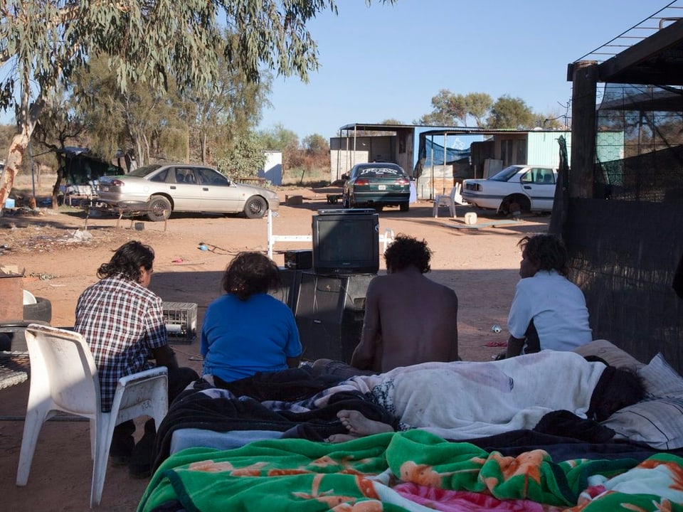 Vier Aborigines sitzen vor einer Wellblechbehausung.