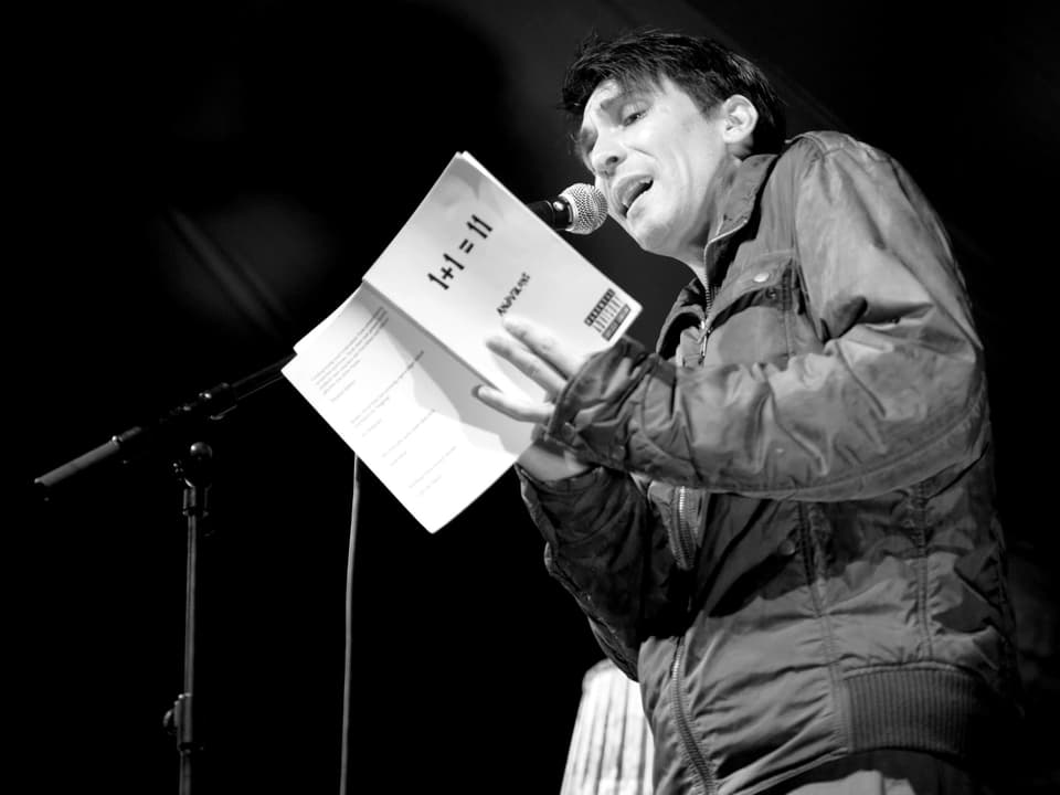 Ein Mann steht auf einer Bühne, hält ein weisses Buch in der Hand und spricht in ein Mirkofon. 