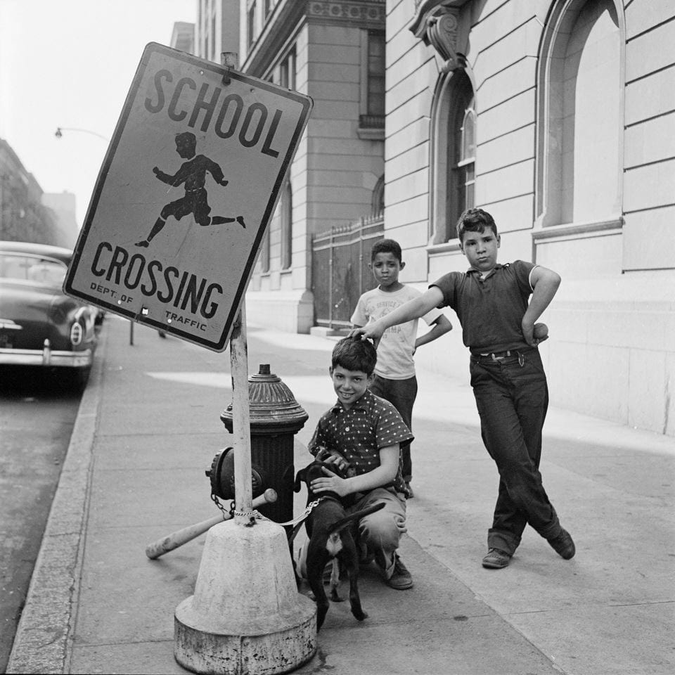 Drei Kinder auf der Strasse. Sie stehen neben einem Schild mit der Aufschrift «School Crossing».