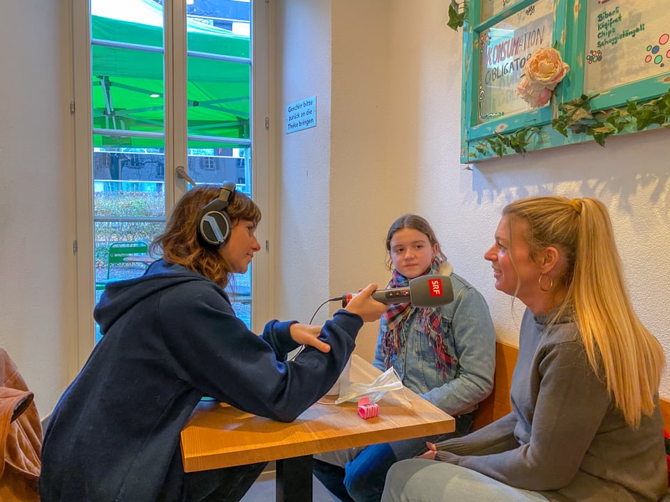 Ein Mädchen und zwei Frauen sitzen in einem Cafe, die blonde Frau spricht ins Mikrofon.
