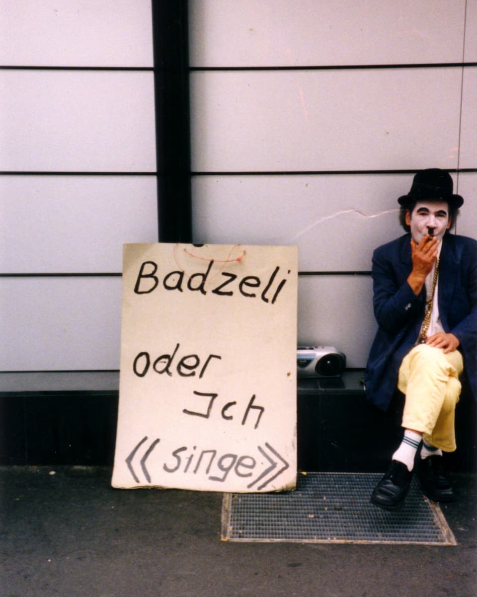 Ein Mann mit Charlie-Chaplin-Verkleidung sitzt neben einem Karton mit der Aufschrift «Badzeli oder ich ‹singe›»