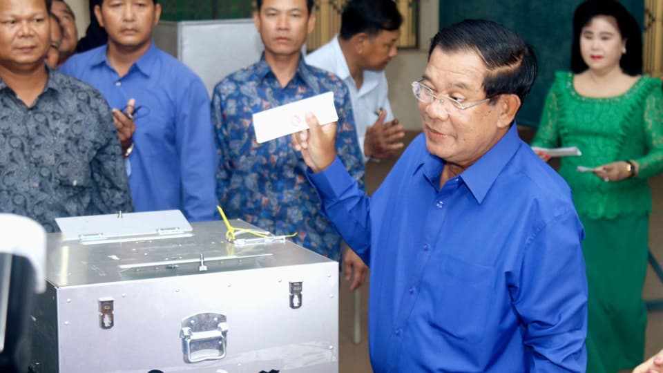 Mann in blauem Hemd und Brille steht vor Wahlurne, Personen rundum schauen ihm zu.