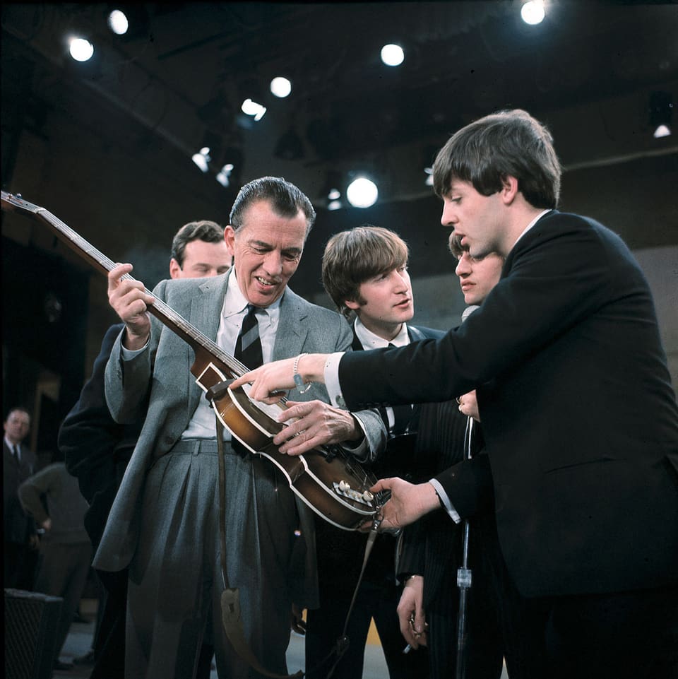 Der TV-Entertainer Es Sullivan hält eine Gitarre, Paul McCartney von den Beatles steht neben ihm.