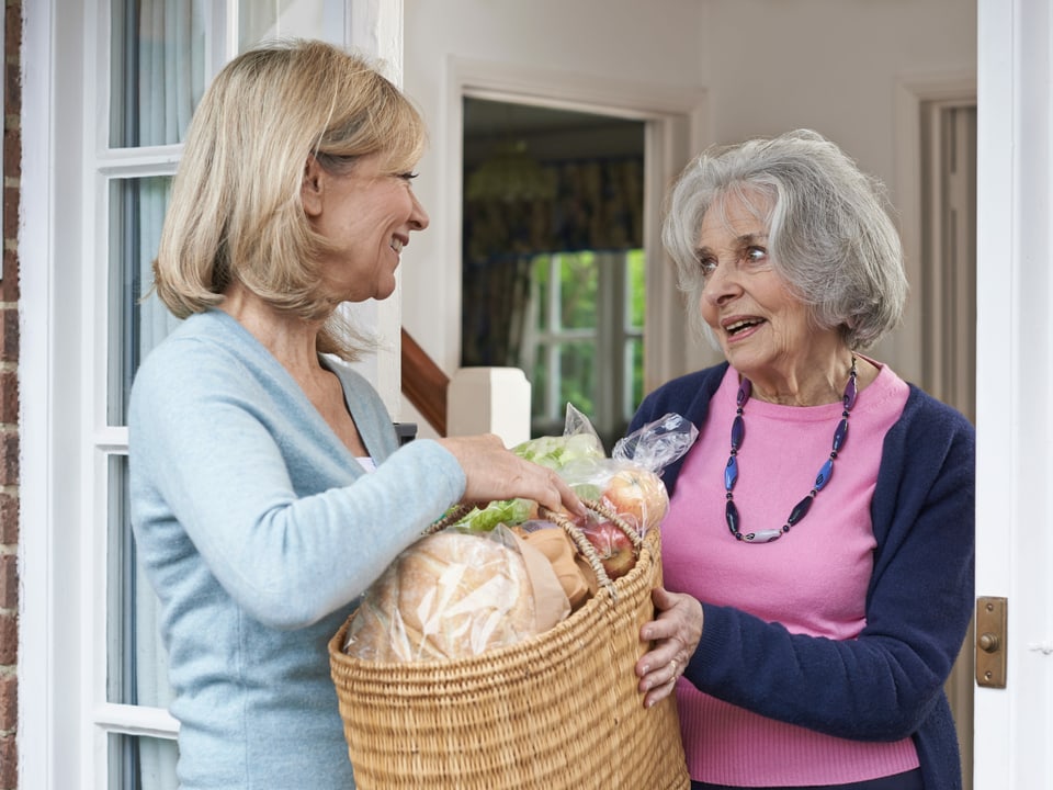 eine Frau übergibt einer Seniorin eine Tasche mit Einkäufen.