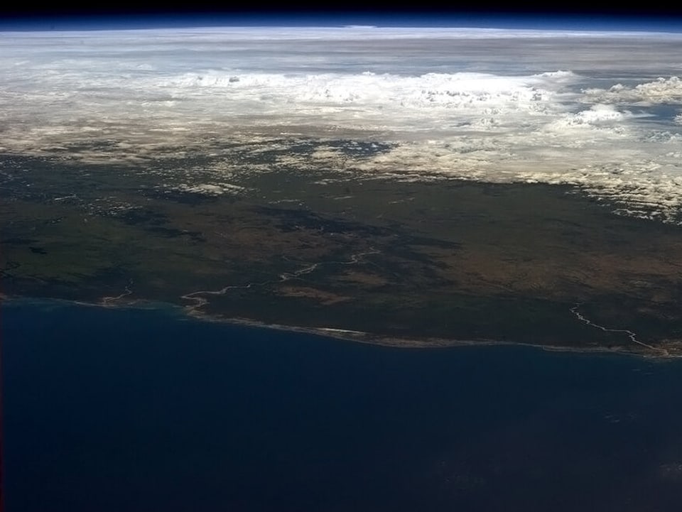 Blick aus dem Orbit auf die wolkenfreie Küste Madagaskars. Im Hintergrund ist ein breites Wolkenband zu sehen.