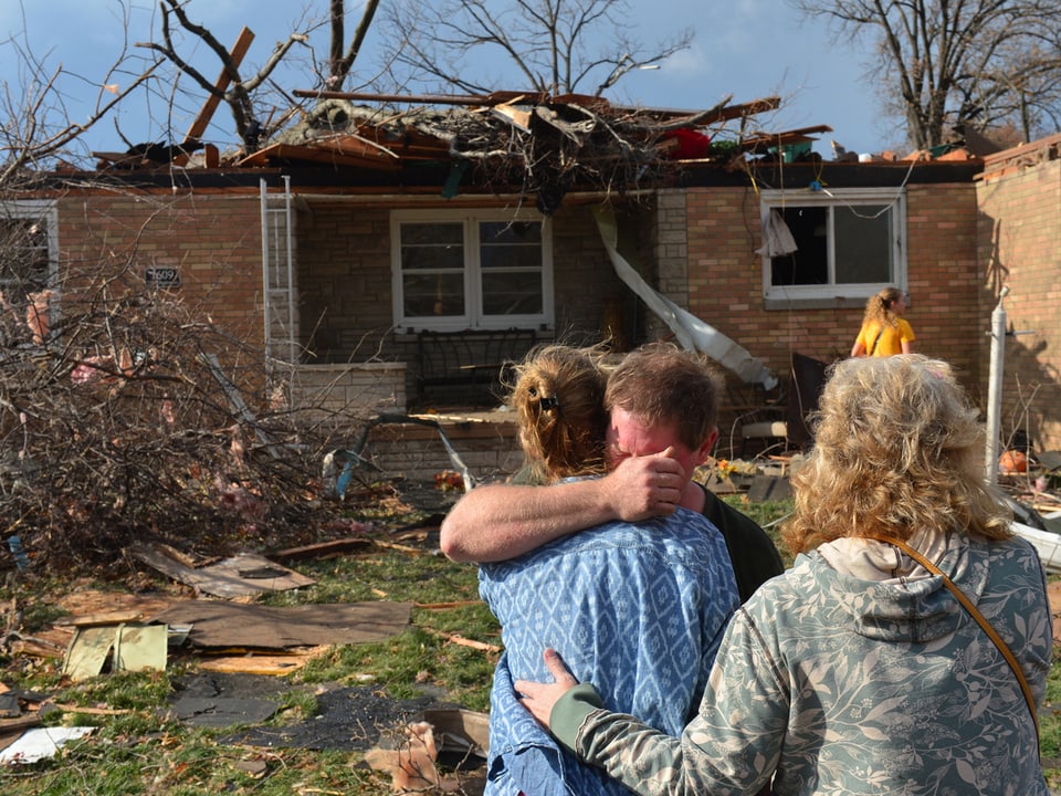 Menschen halten sich in den Armen vor einem zerstörten Haus.