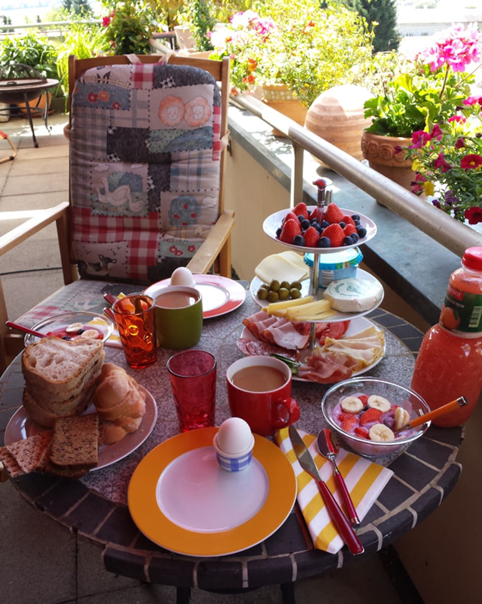 Frühstückstisch auf dem Balkon. Brot, Ei, Kaffe, Früchte, Käse, Fleisch, Müesli, Orangensaft.