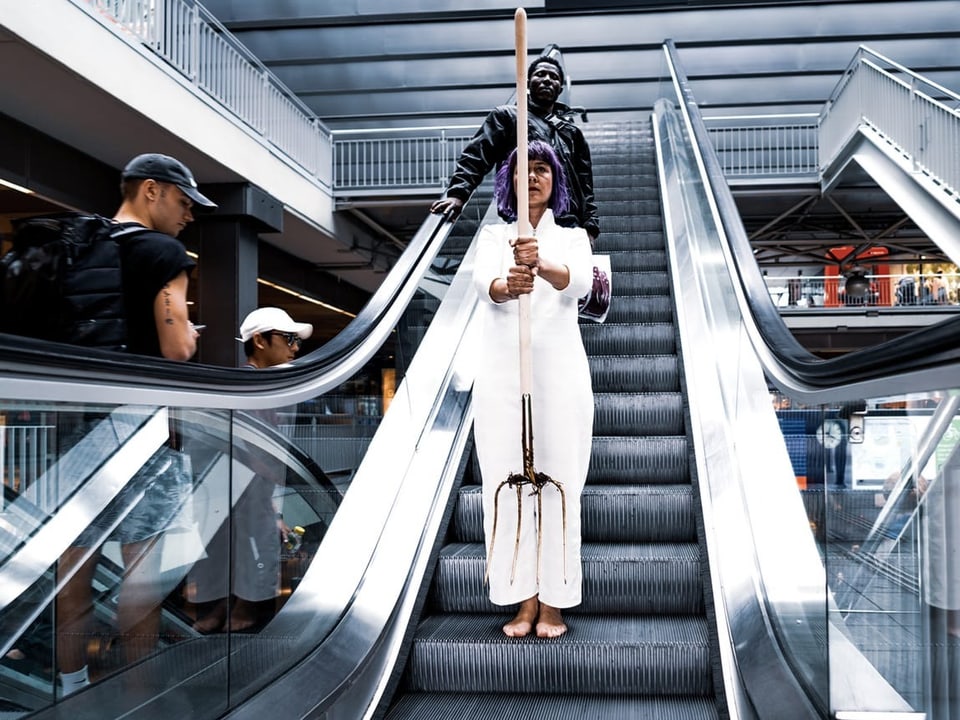 Frau im weissen Anzug steht mit Mistgabel auf einer Rolltreppe
