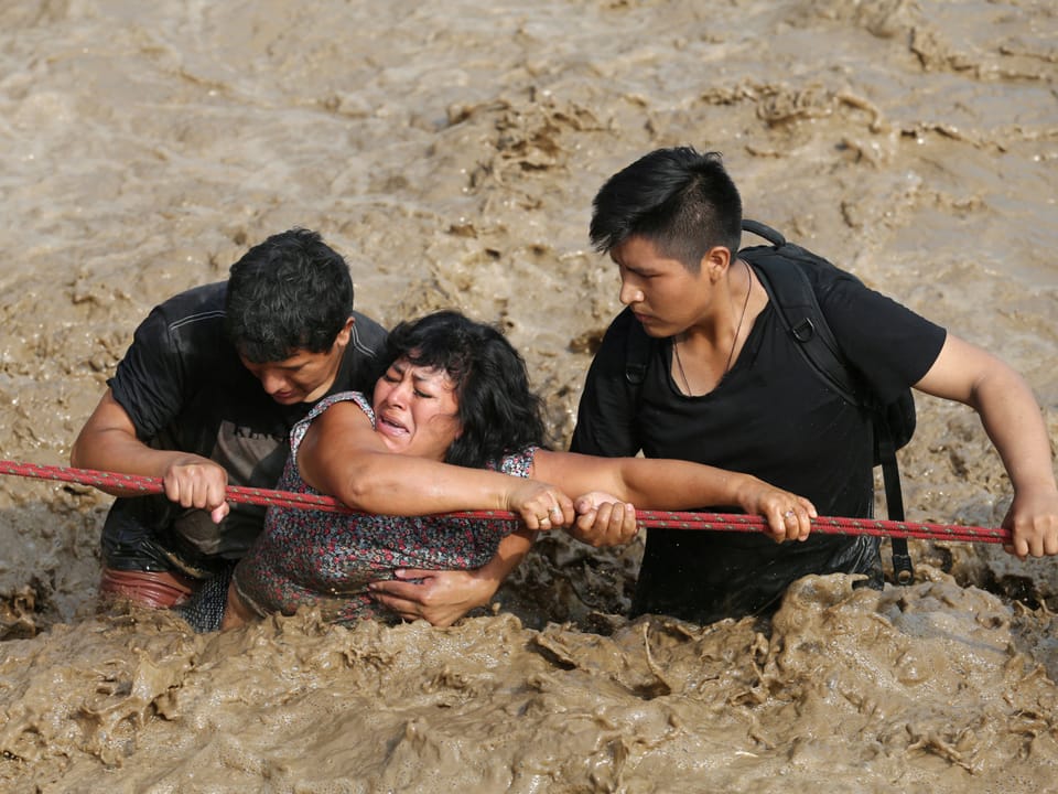 Drei Leute kämpfen sich mit Seil durch den überschwemmten Fluss.
