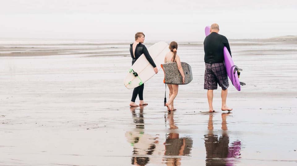 Junge Kinder und ein älterer Herr mit Surfbrett unterm Arm am Strand