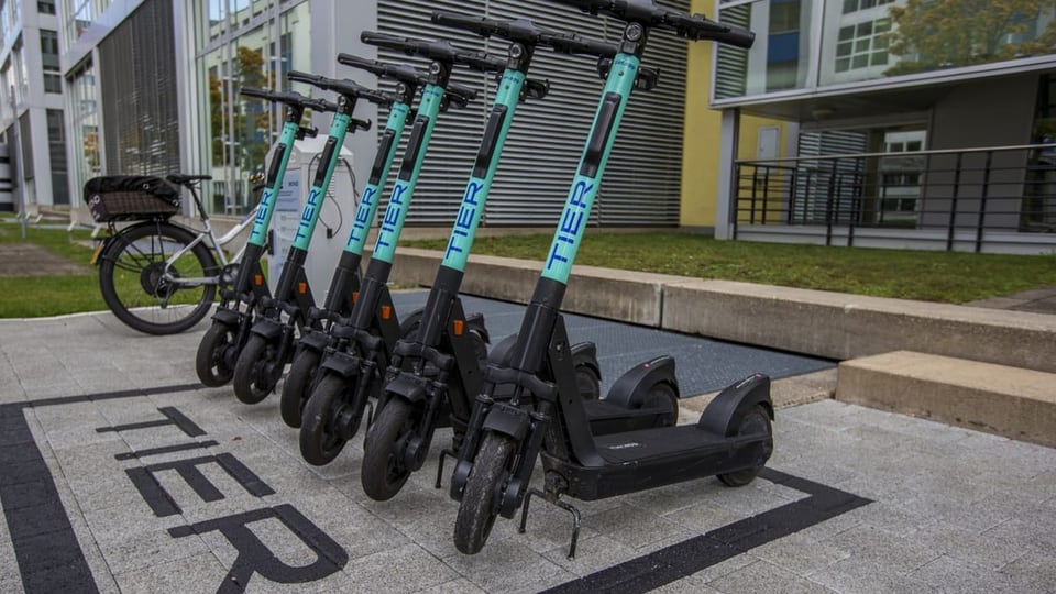 Sechs nebeneinander aufgereihte E-Trottis stehen auf einem Parkplatz