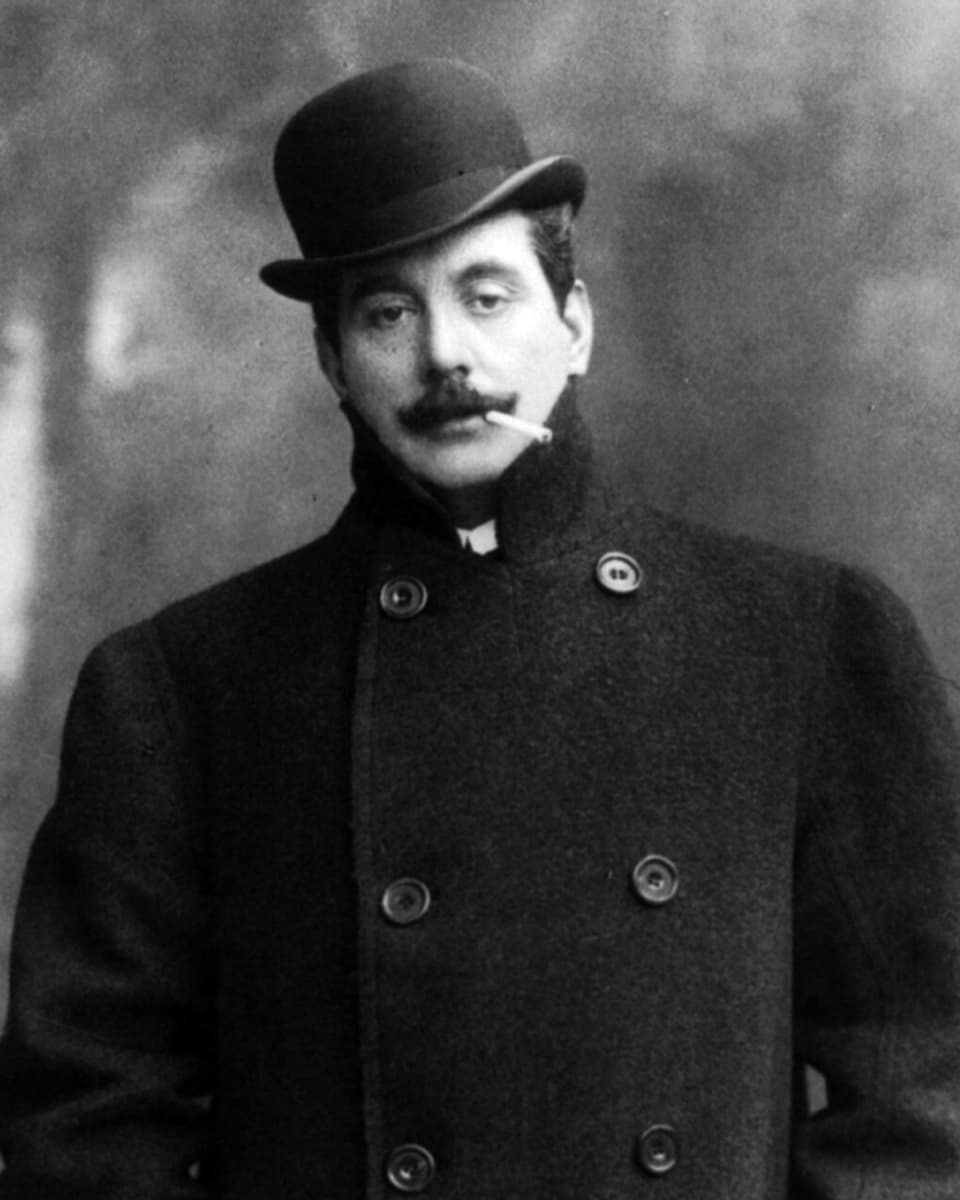 Puccini mit Mantel, Hut und Zigarette.