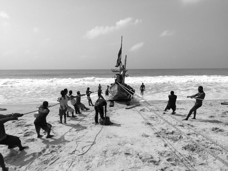 Schwarz-Weiss-Aufnahme: Rund ein Dutzend Männer ziehen das Boot an rund sieben langen Seilen durch den Sand.
