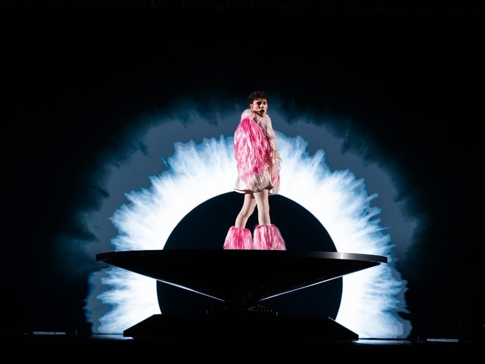 Person in rosa Federkleidung steht auf einer Bühne vor einem leuchtenden Kreishintergrund.