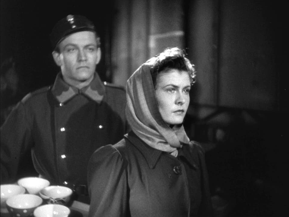 Eine Frau (Gilberte) blickt nüchtern in die Nacht hinaus. Hinter ihr steht ein Junger Soldat.