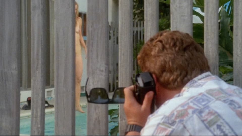 Man looks voyeuristically through a picket fence at a naked woman climbing into the pool.