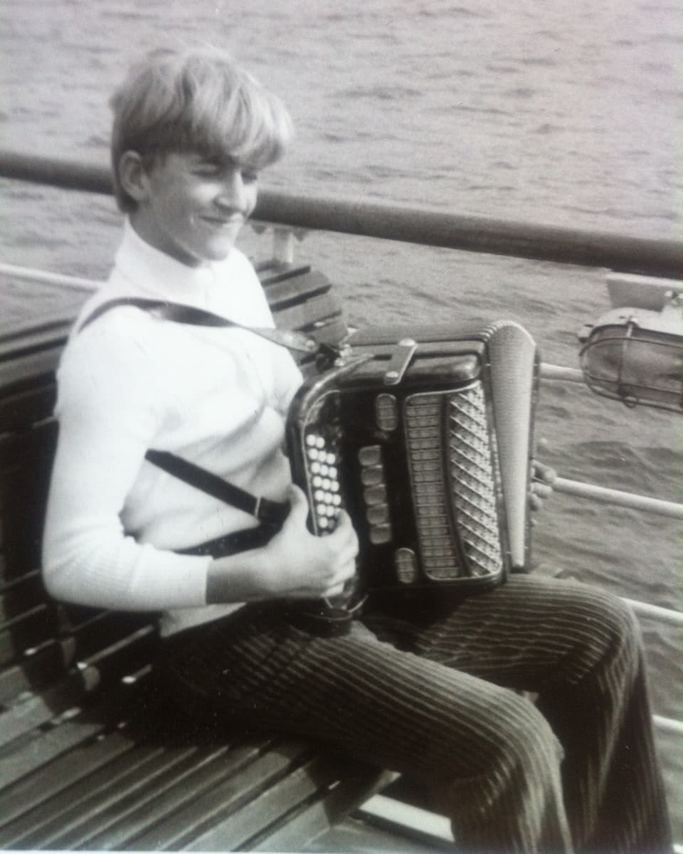 Ein junge Mann sitzt mit einem Akkordeon auf dem Deck eines Schiffes.