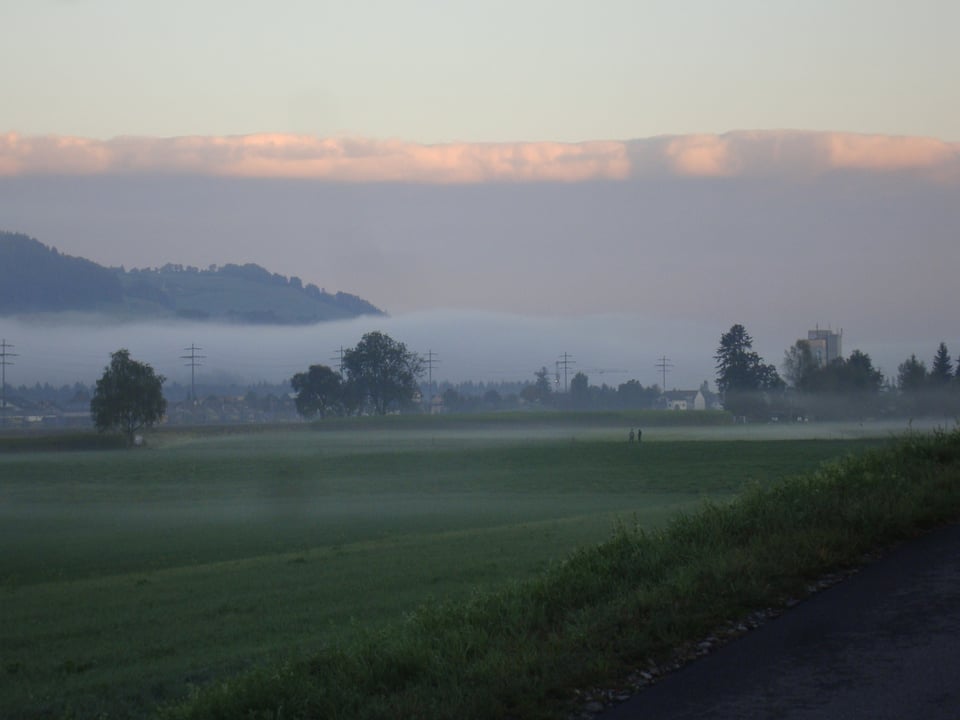 Im Vordergrund ein grünes Tal mit einer Nebelbank am Fusse eines Hügels. Darüber eine dünne Hochnebeldecke, die von der Sonne beschonnen wird.