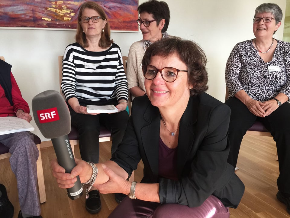 Ursula Schuppisser kniet mit Mikrofon im Kreis, in dem Leute sitzen.